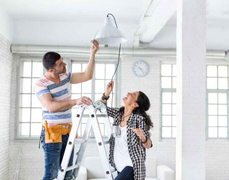 a woman in a white shirt and a handyman in a ladder changing electrical of a lamp